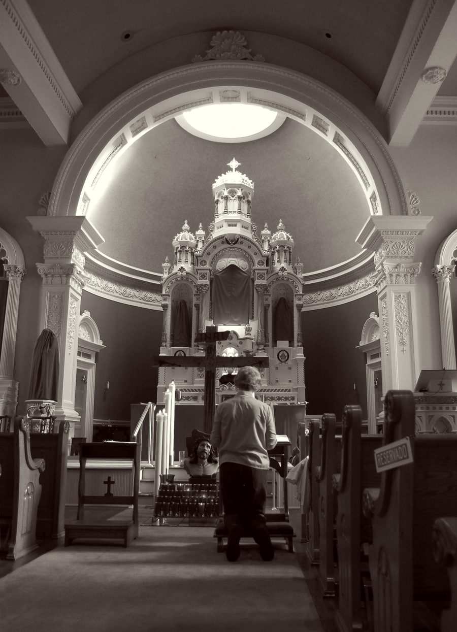 A black-and-white photograph of a parishioner kneeling in prayer before the altar of Seelos Church on Good Friday