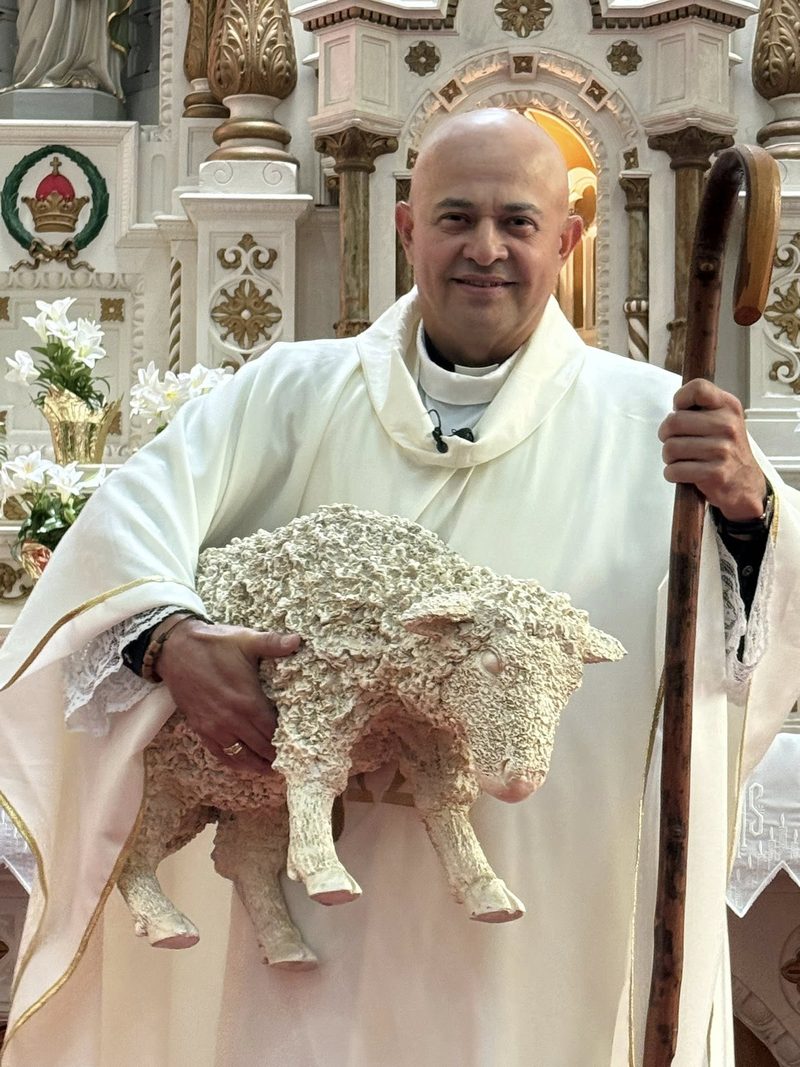 Father Alex Guzmán in white vestments, holding a sculpted lamb and a shepherd&rsquo;s crook, smiling at the camera in the sanctuary